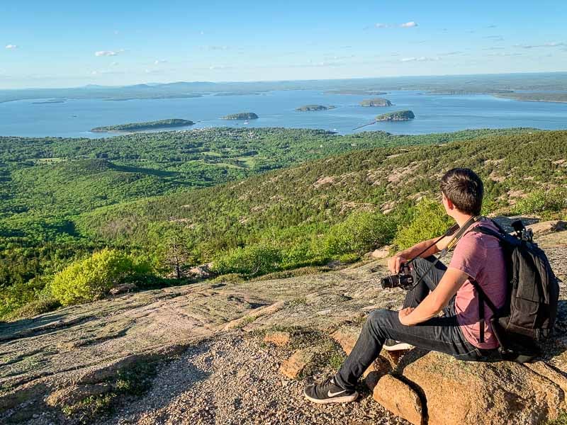 A man sitting on a rock, gazing at a serene lake surrounded by nature's beauty in Cadillac Mountain