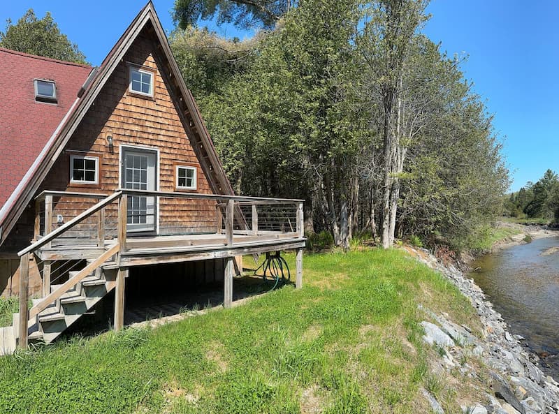 A wooden A-frame cabin with a deck, situated beside a river and surrounded by trees.