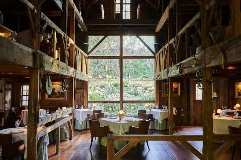 The elegant interior of a renovated barn restaurant with high ceilings and a large window.